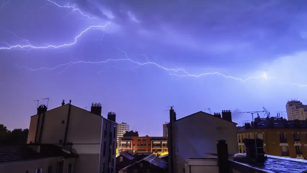 Vue nocturne de toits urbains sous un ciel zébré par des éclairs impressionnants.