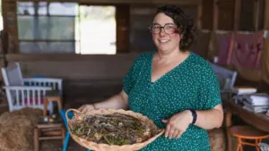 Une femme souriante aux cheveux courts porte une robe verte, tenant un plateau de plantes.