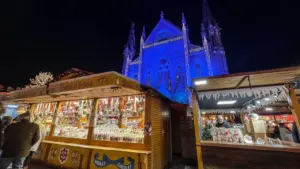 Des étals de marché illuminés devant une église bleue sous un ciel nocturne.