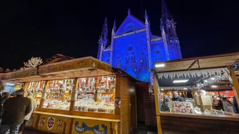 Des étals de marché illuminés devant une église bleue sous un ciel nocturne.
