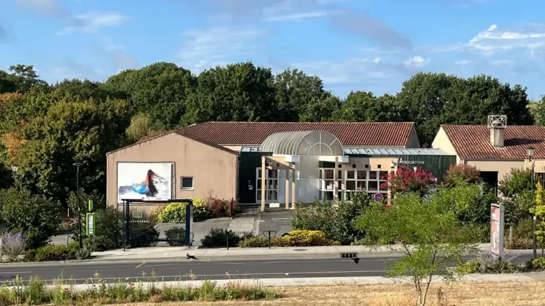 Bâtiment moderne avec toit en tuiles et entrée vitrée entouré d'arbres sous un ciel bleu.