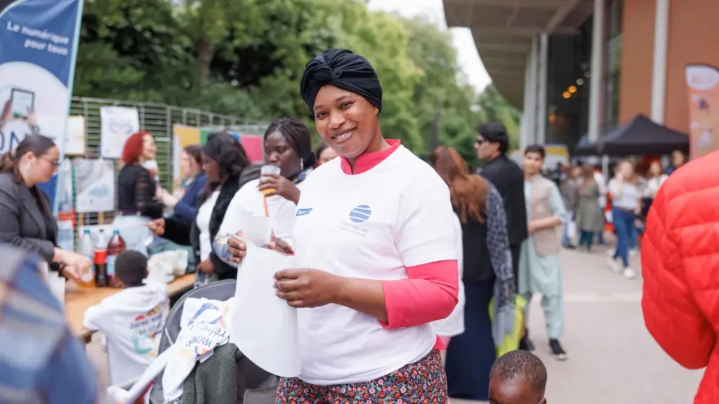 Une femme souriante portant un turban noir et un t-shirt blanc lors d'un événement extérieur.