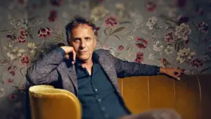 Un homme d'âge mûr pose assis sur un canapé jaune devant un papier peint fleuri.
