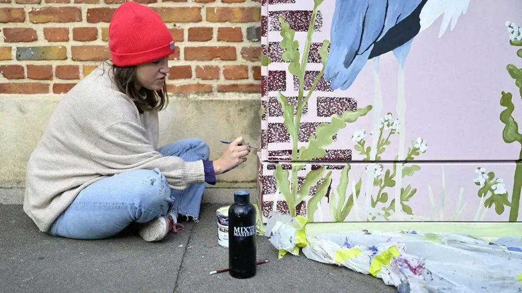 Une femme avec un bonnet rouge peint une fresque colorée sur un mur extérieur.